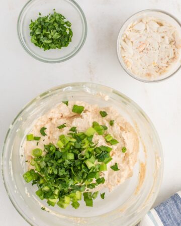 Cream cheese and sour cream with chopped green onions to make crab dip in a glass bowl.