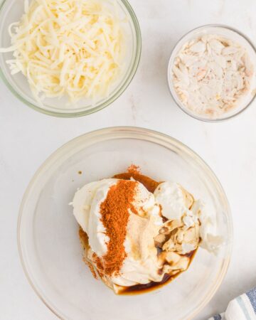 Hot crab dip ingredients including old bay seasoning and cream cheese in a glass bowl.