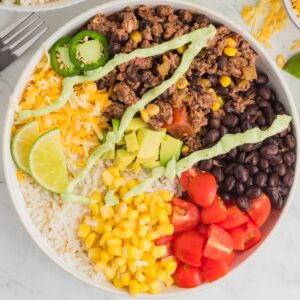 A beef burrito bowl with rice, corn, tomatoes, beans, and shredded cheese with an avocado crema.