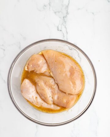 A glass bowl with chicken in a Greek marinade to make Greek Chicken Salad. 