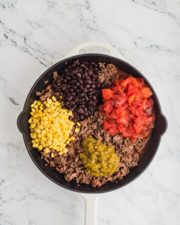 A cast iron pan with browned ground beef and the addition of corn, beans, green chilies, and tomatoes. 
