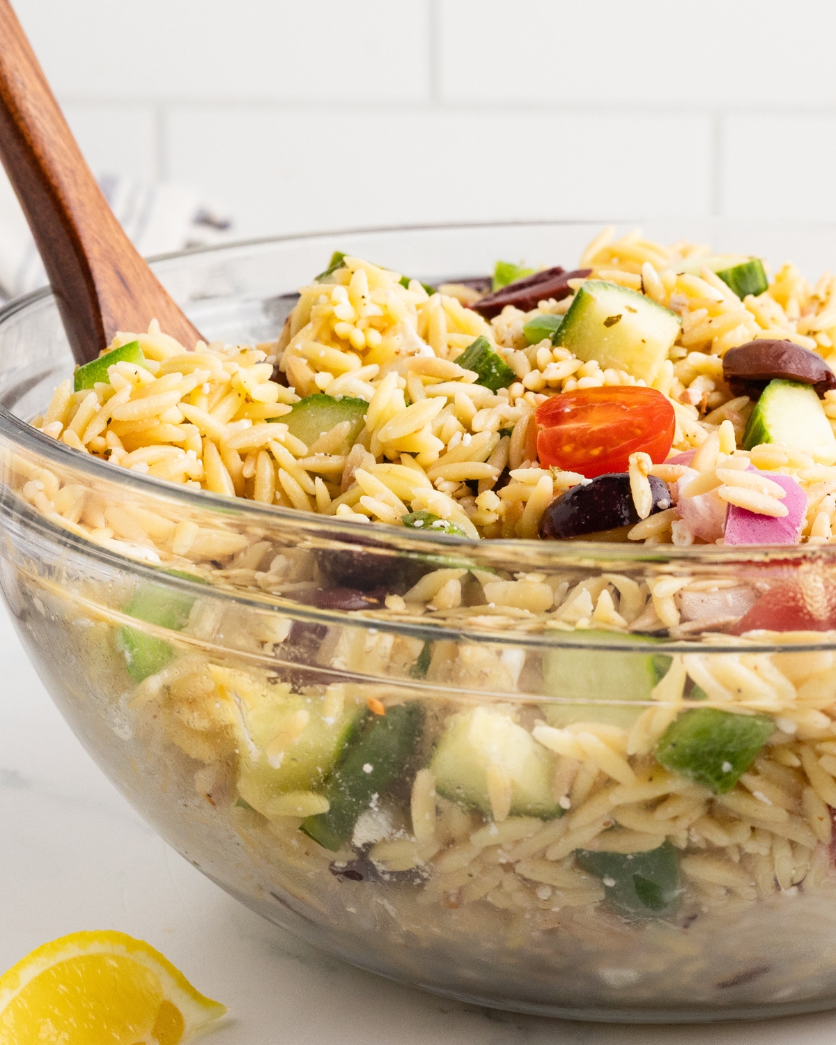 A glass bowl with cooked orzo and tomatoes, cucumbers, and feta.