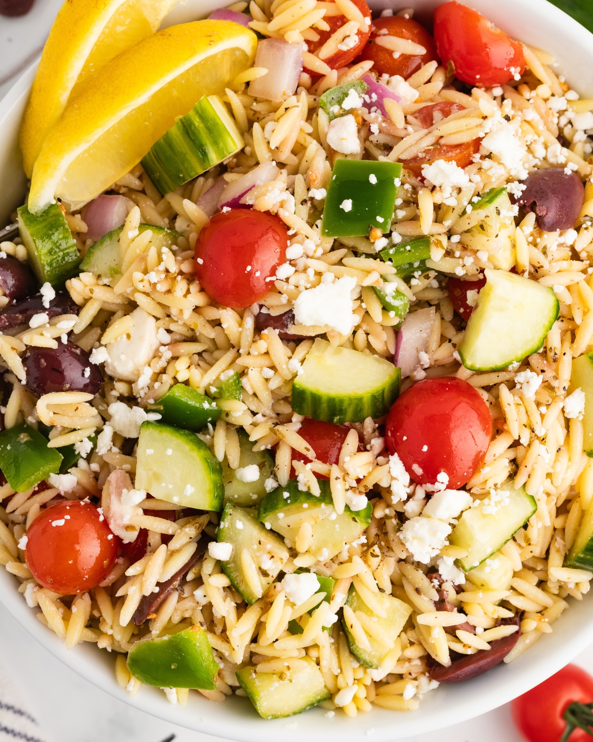 An orzo salad with tomatoes and cucumber in a white bowl with sliced lemon and feta.