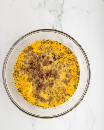 A glass bowl with whisked egg, dried herbs, seasonings, and crumbled breakfast pork sausage.