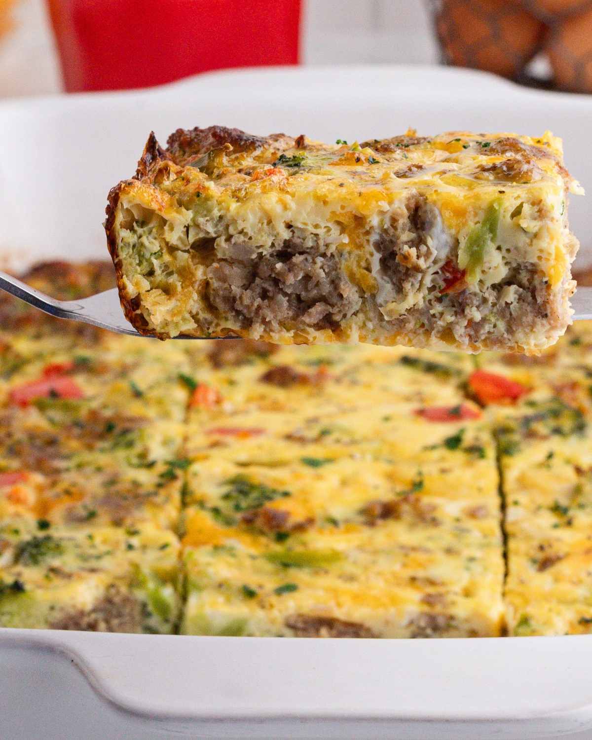 A wedge of a breakfast casserole made with eggs, ground pork sausage, broccoli, red peppers, and shredded cheddar cheese.
